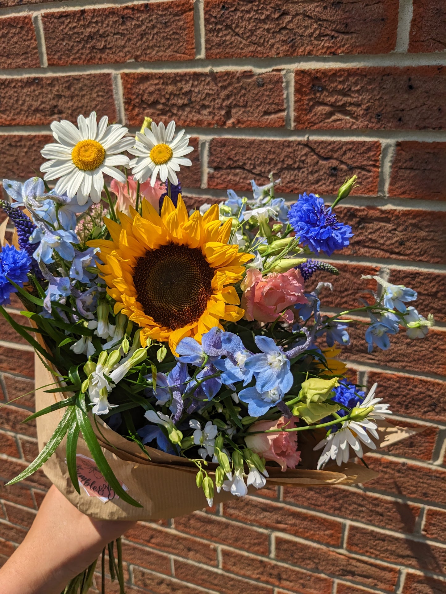 a bright bunch of flowers with sunflowers daisies delphiniums coral lizzi wrapped in brown paper with open stem